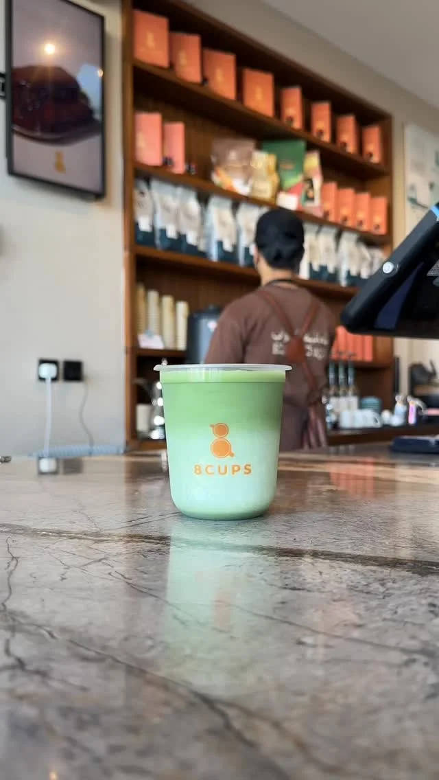 كوب 8CUPS شفاف من الماتشا على رخام الكاونتر، وخلفه أكياس القهوة الطينية